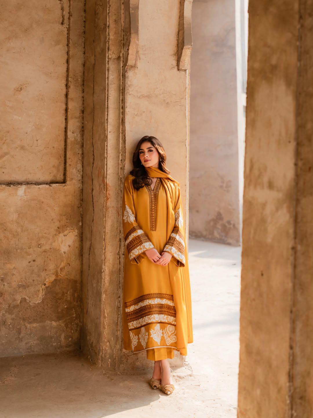 Woman in a yellow traditional outfit standing in a sunlit stone corridor. House Of Beenora | Best women clothing brand in Pakistan