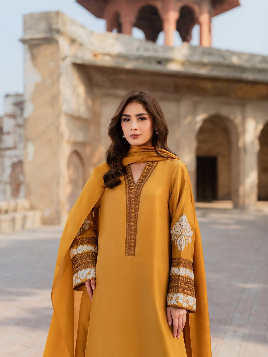 Woman in a mustard yellow dress with intricate patterns standing in front of historical architecture.