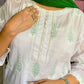Person wearing a white embroidered top with green scarf, standing against a floral curtain background.