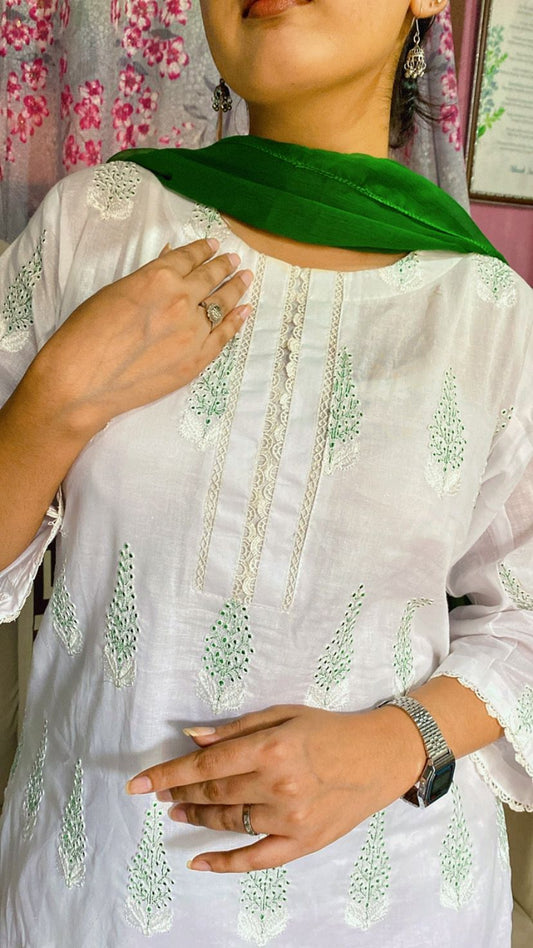 Person wearing a white embroidered top with green scarf, standing against a floral curtain background.