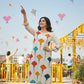 Woman in a colorful dress with geometric pattern standing outdoors with kites flying around her.