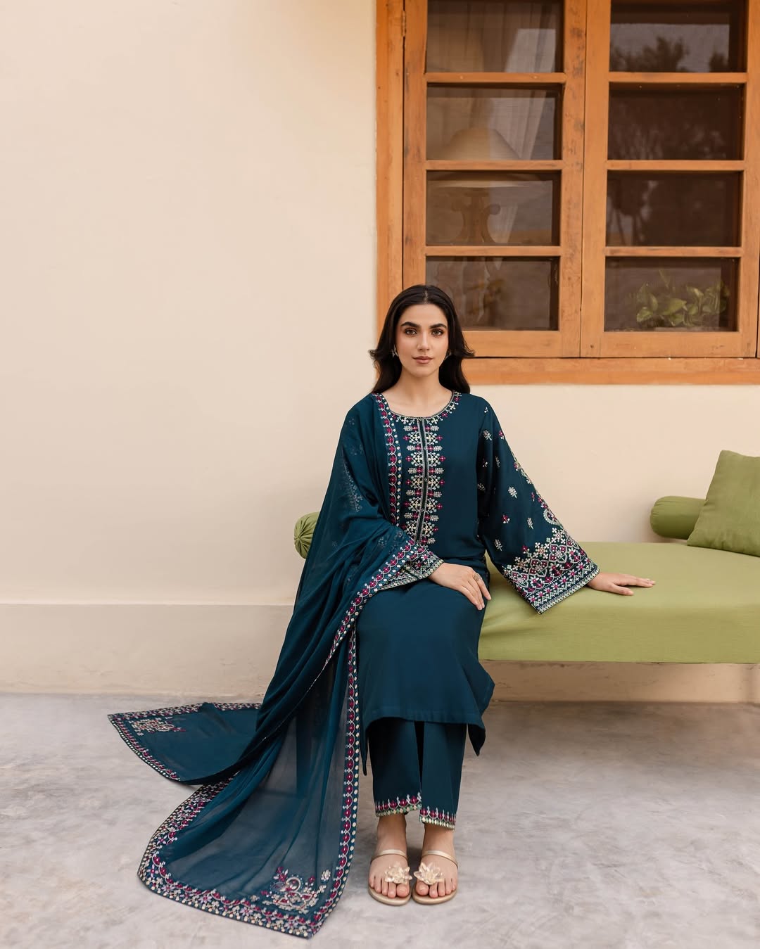 Woman in a blue traditional outfit sitting on a green couch in a room with a wooden window.