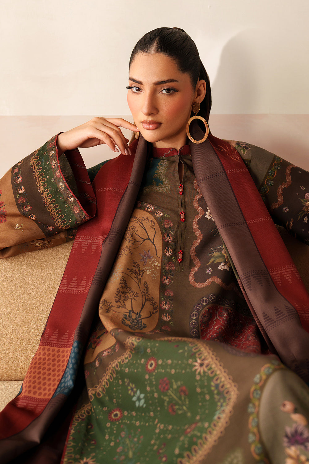 Woman wearing a traditional embroidered outfit with a red dupatta, sitting on a beige couch.